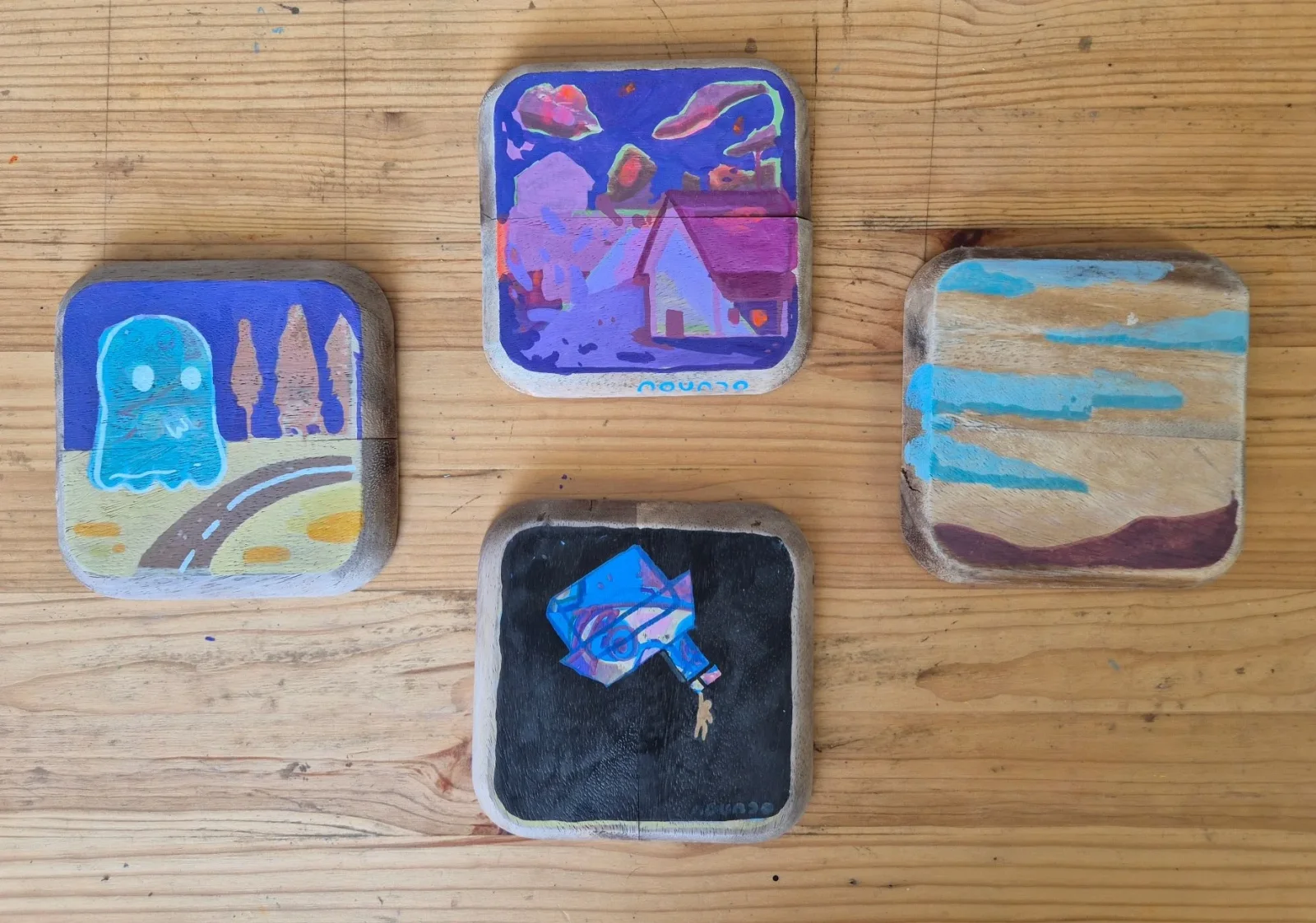 Four small square painted plates: a blue ghost by a road, a purple house under a night sky, a black plate with a blue figure,
